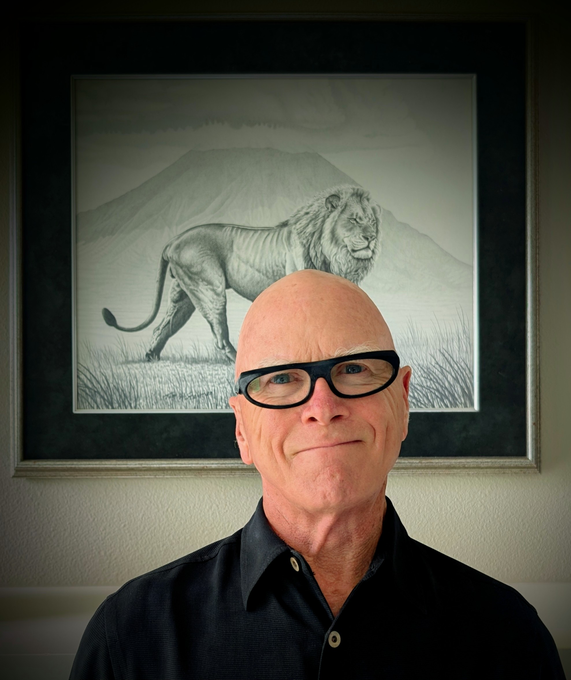 A 2025 portrait of Bill Perkins — round warm face, thick horn-rimmed glasses, a soft smile, photographed in front of a framed lion sketch